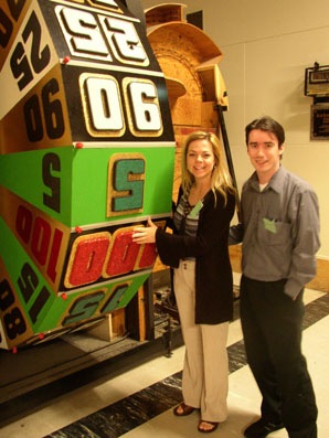 Two individuals standing next to a large wheel with numbers on it, likely from a game show setting.