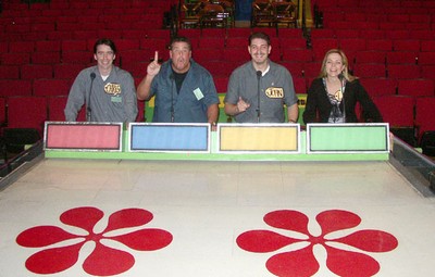 Four individuals standing behind a game show podium with colorful panels and flower designs on the ground, set in a backstage environment.