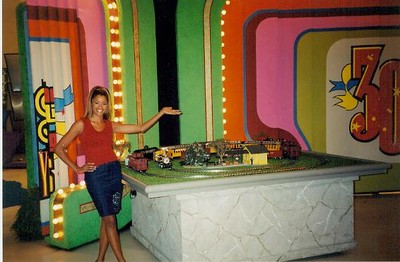 A woman stands next to a miniature train set displayed on a table, with colorful game show backdrops behind her.