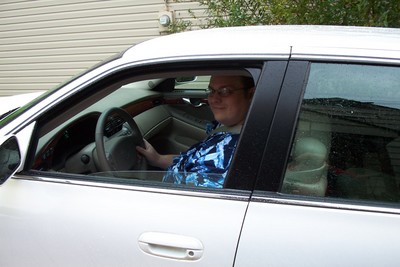 Travis Schario wearing a blue floral shirt is sitting in the driver's seat of a silver car, smiling at the camera.