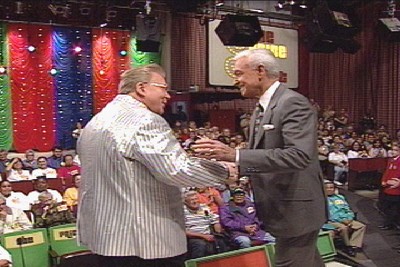 Two men interacting on stage at The Price is Right, surrounded by audience members and colorful backdrop decorations.