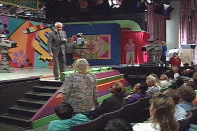 Bob Barker speaking to an audience on the set of The Price is Right, with colorful decorations and video cameras in the background.