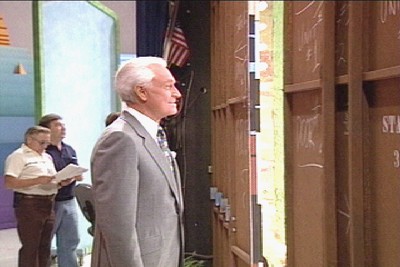 Bob Barker in a gray suit looking at a large set door with chalk markings during a production of a game show.