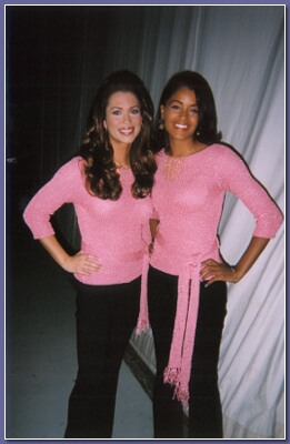 Claudia Jordan and Brandi Sherwood in matching pink sweaters and black pants standing side by side, smiling. The background is a light-colored curtain.