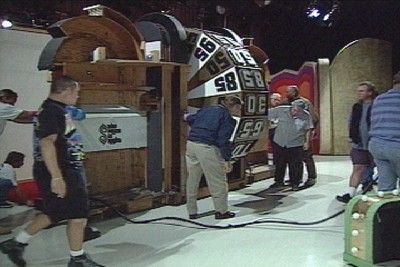 A behind-the-scenes view of the set featuring crew members moving the Big Wheel into position, with various studio equipment present.