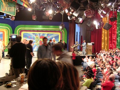 A busy television studio set for a game show, with colorful backdrops and bright stage lighting. Crew members are setting up while an audience watches eagerly from their seats.