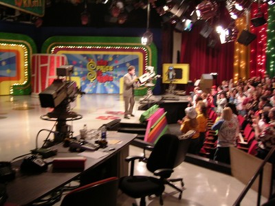 A blurred photo of a game show set where a host stands in front of an audience, with colorful backdrops and studio lights.