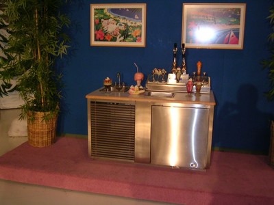 A retro-style bar counter featuring a variety of drinks and dessert toppings, set against a blue backdrop with framed pictures and greenery on either side.
