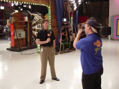 A television studio scene featuring two men, one standing and speaking into a microphone while the other is filming with a camera, against a backdrop of stage equipment.