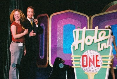 A woman holds a microphone and stands next to a host on stage, with a brightly colored 'Hole in One' sign in the background.