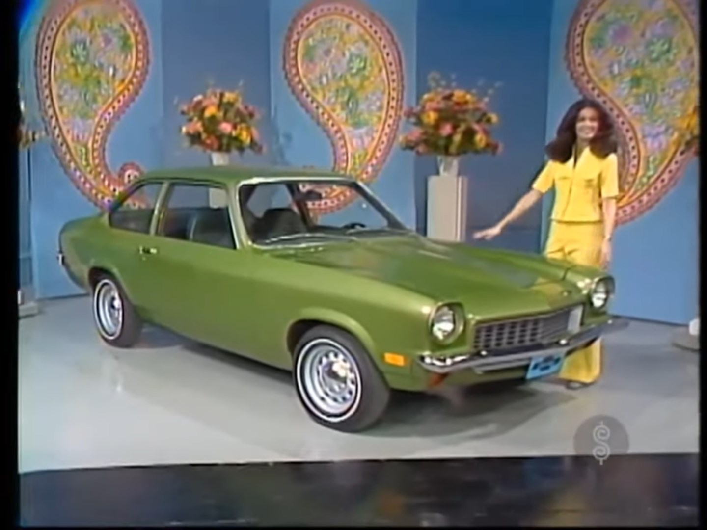 A woman in a yellow outfit stands next to a green 1973 Chevy Vega on a colorful stage backdrop decorated with flowers.