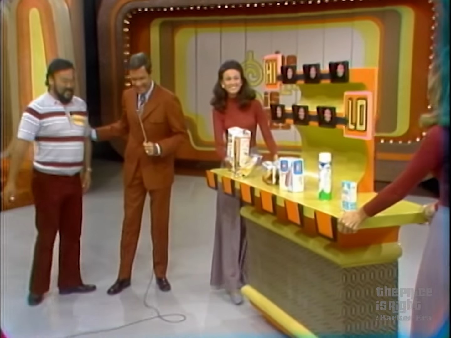 A contestant interacts with the host on stage during the Hi Lo game from The Price is Right, with grocery products displayed on a colorful game board.