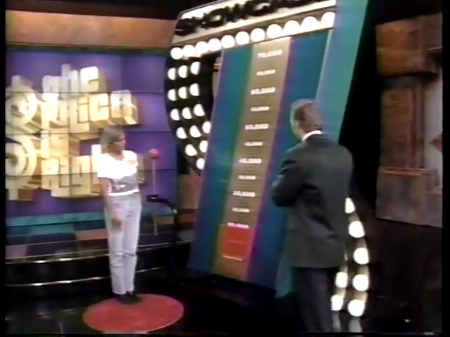 A contestant is playing the Range Game on The Price is Right, holding a button to stop the moving range finder on a colorful scale displayed behind them.