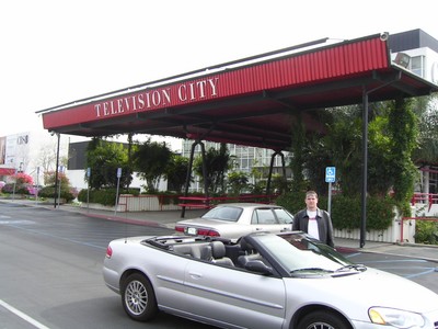 Exterior view of Television City with a person standing beside a silver convertible car.