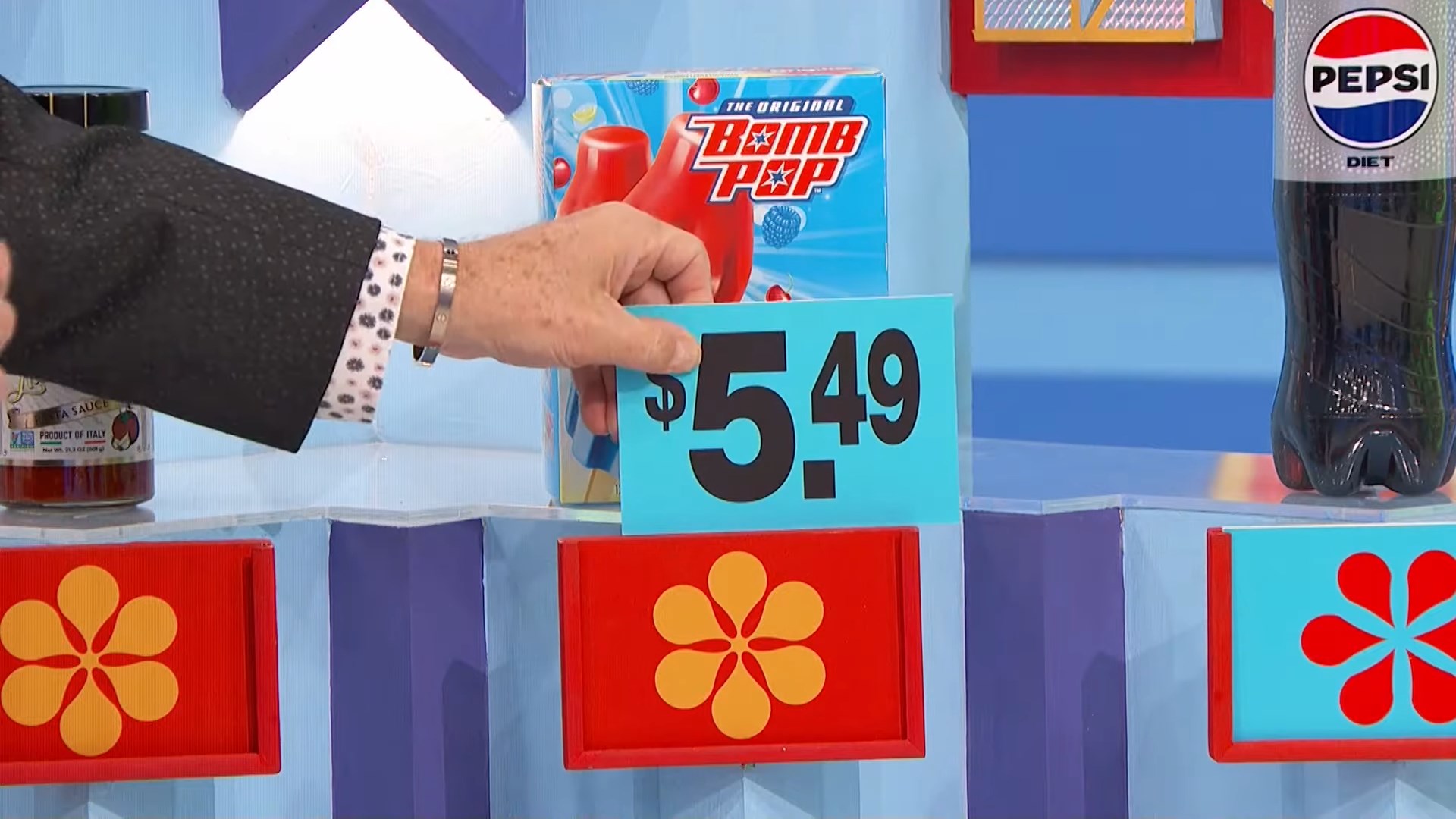 A contestant's hand reveals a price tag of .49 for a box of Bomb Pop treats on a colorful game show set.