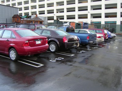 A row of parked vehicles with various colors, including a red car and a dark-colored car, beside a building with a multi-story parking structure in the background.