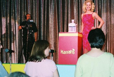 A woman in a pink dress stands on stage next to a podium with the Harrah's logo, smiling at the audience. A camera operator is in the background capturing the moment.