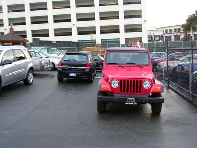 A red Jeep parked in a lot, with several other vehicles in the background and a multi-story building partially visible.