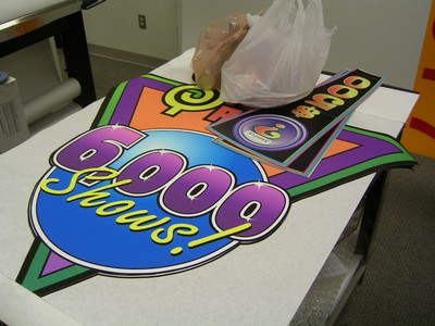 A large colorful sign displaying '6000 Shows!' on a table, alongside some promotional materials and a brown bag.