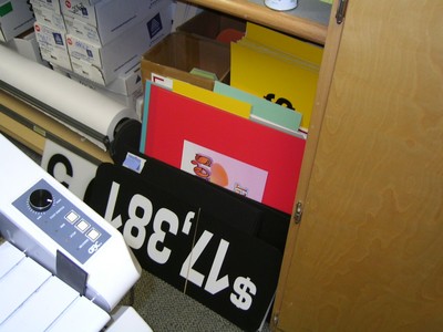 A collection of colorful signs and papers stored in a cabinet, featuring a prominent black sign with white text, alongside various other colored sheets.