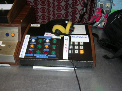 Close-up of a studio control panel with buttons labeled 'Miami' and 'Scottsdale' and a pair of yellow headphones, controlling the Bargain Game.