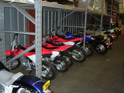 A row of Honda dirt bikes in a storage area with metal shelving.