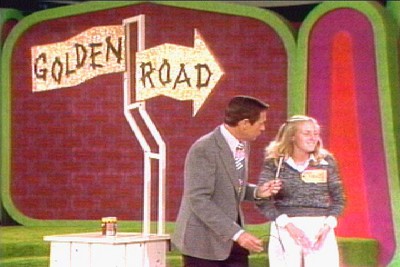 A host and a contestant on the set of The Price is Right stand beside a sign indicating 'Golden Road' in a colorful game show studio.