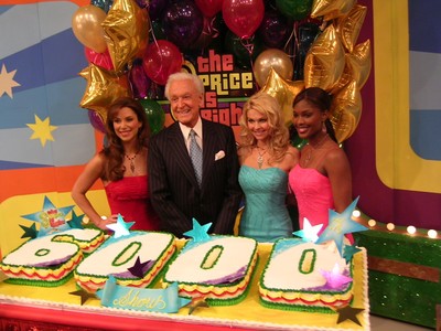A celebration scene from 'The Price is Right' featuring Bob Barker and three models standing behind a large cake decorated with the number 6000 and colorful stars.