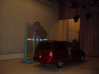 A red minivan parked on a stage in front of a colorful game show backdrop featuring large circular shapes.
