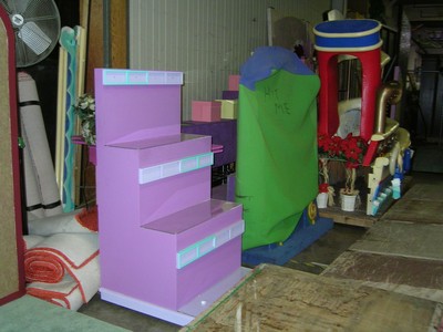 A purple display stand with multiple shelves is positioned in a backstage area, surrounded by various colorful props.