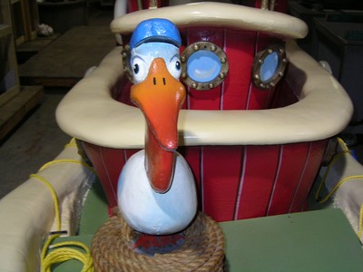 Close-up of a whimsical duck figure wearing a blue cap, positioned at the bow of a red boat with porthole windows.