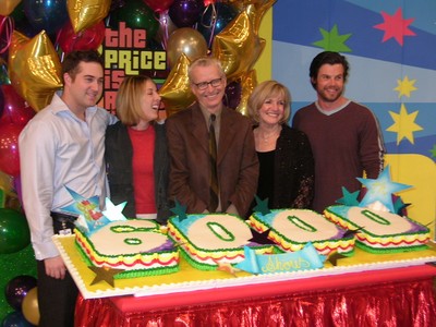 A group of five people celebrating in front of a large cake with the number '8000' on it, decorated with colorful stars and surrounded by balloons.