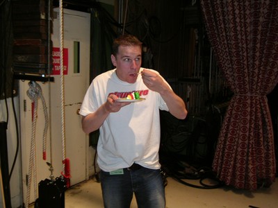 A person in a white shirt holding a plate of colorful dessert while standing in a studio setting with curtains in the background.