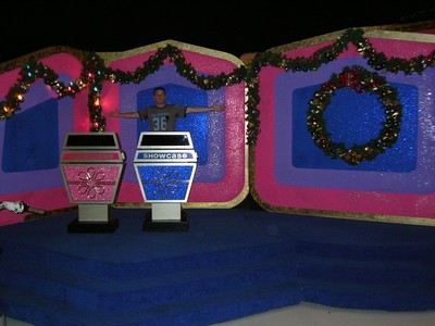 A person stands in front of a colorful game show set featuring two showcase displays, decorated with holiday garlands and ornaments.