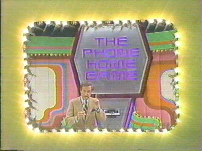 A game show host stands in front of a colorful game setup featuring the words 'THE PHONE HOME GAME' illuminated by lights.