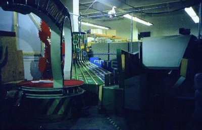 An interior view of the Penny Ante game set, showing the colorful game board and various equipment in a warehouse. Items include the large circular podium and green ramps typically used in the game.