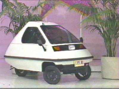 A small, futuristic-looking white Zoe Zipper car with a unique shape, parked on a stage decorated with palm plants.
