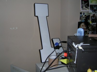An illuminated letter 'L' sign on a wall next to a computer setup, with some household items visible in the background.