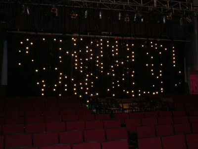 A dimly lit stage with a backdrop of hanging lights and empty red theater seats in the foreground.