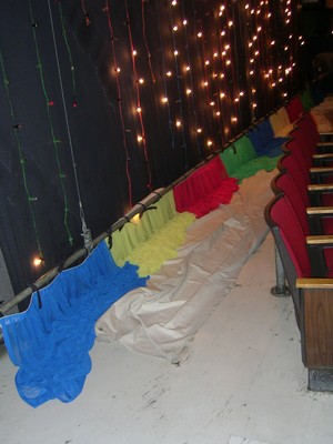 Colorful curtains draped along a studio wall with twinkling lights in the background and red theater seats visible in the foreground.