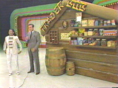 Bob Barker and a contestant on a game show set standing next to a wooden structure labeled 'Trader Bob' filled with various packaged goods.