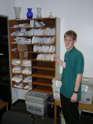 Scott Robinson standing next to a wooden shelf filled with organized file folders, with a box for mail and a table beside it.