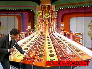 A game show set for Penny Ante featuring colorful prices and oversized icons, with a host demonstrating a contestant's guess.