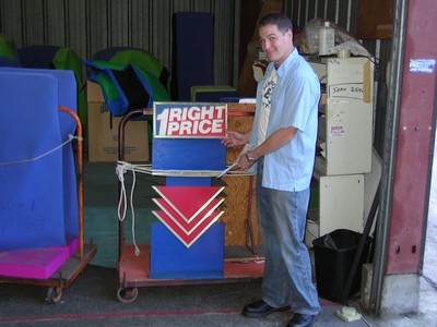 John Sly stands next to a colorful game display labeled '1 RIGHT PRICE' in a storage area filled with game show props.