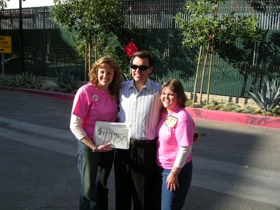 Three people smiling in a parking lot, with two wearing pink shirts and holding a sign that reads $77,750.