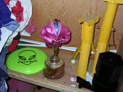A shelf cluttered with a pink suit trophy, a green frisbee with an alien design, and various tools.