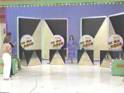 A scene from 'The Price is Right' showing  three large game show panels marked 'on the nose' with a host standing in front and a contestant looking on from the side.
