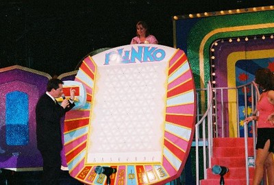 A host presenting the Plinko game with a large Plinko board on stage, featuring two contestants and colorful set pieces in the background.