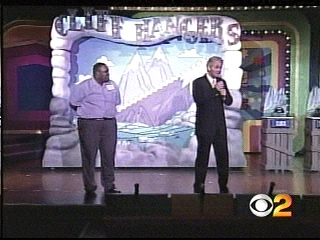 Two hosts standing on stage at a game show, speaking into microphones, with a colorful backdrop featuring mountains.