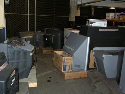 A collection of old televisions stored in a dimly lit area, some placed on wooden pallets and boxes.